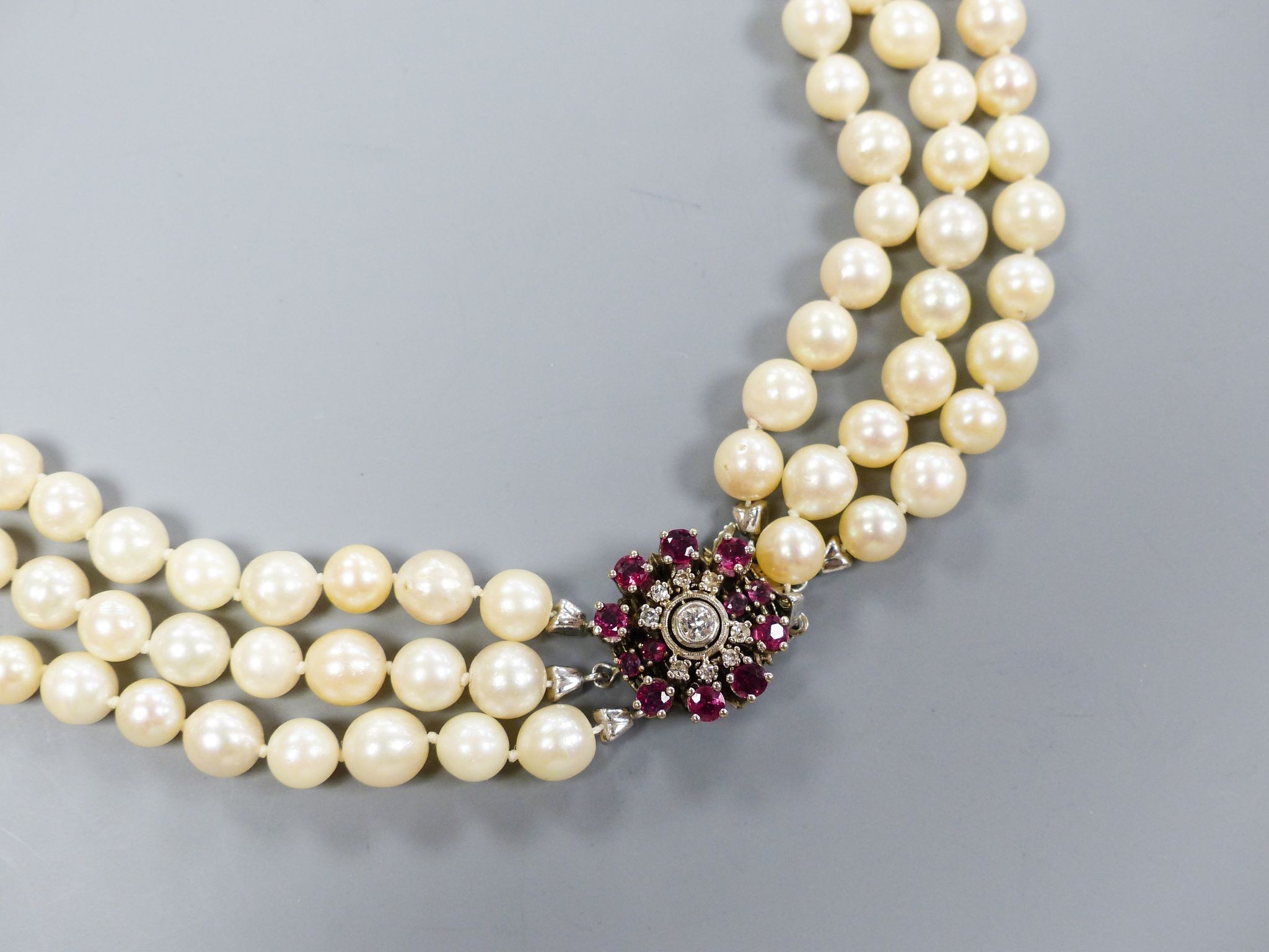 A triple strand cultured pearl necklace, with 585 white metal, ruby and diamond cluster set central motif and 585 white metal clasp, approx. 48cm, gross weight 104.6 grams